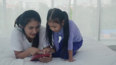 Genç Asyalı anne ve küçük kızı birlikte tablet bilgisayara bakıyor. Yatak odasında öğrenme ve eğitim için. Anne ve kız evde internet, aile ve teknoloji konsepti için tablet izliyor..