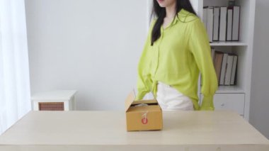 Genç Asyalı bir kadın evdeyken online alışveriş ile paket kutusunu açıp açıyor. İnternetten alışveriş yapan bir kadın. Heyecanlı ve teslimat konseptiyle bakıyor..
