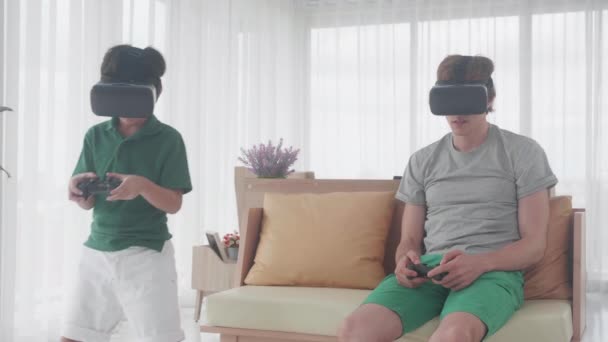 年轻的亚洲父亲和儿子坐在家里沙发上玩电子游戏的虚拟现实 男人和男孩一起做沙发上放松的活动爱好 家庭观念 — 图库视频影像