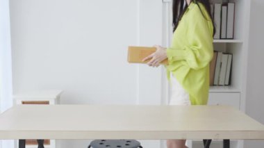 Genç Asyalı bir kadın elinde karton kutuyla eve gidiyor, mutlu bir kadın koli kutusu taşıyor ve onu masanın üstüne koyup hediye paketi, hediye paketi, online alışveriş mağazası ve servis konseptine bakıyor..