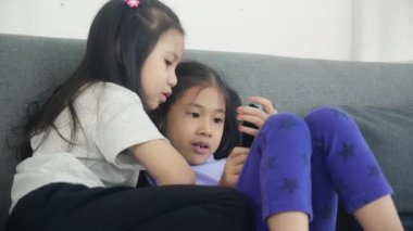 Mutlu Asyalı küçük çocuk ve arkadaşları evdeki kanepede akıllı telefon oynuyorlar, çocuklar koltukta birlikte telefon kullanıyorlar, kız eğlence, yaşam tarzı ve iletişim konsepti için akıllı telefon izliyor..
