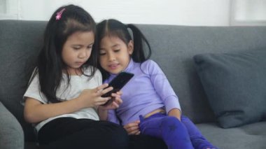 Mutlu Asyalı küçük çocuk ve arkadaşları evdeki kanepede akıllı telefon oynuyorlar, çocuklar koltukta birlikte telefon kullanıyorlar, kız eğlence, yaşam tarzı ve iletişim konsepti için akıllı telefon izliyor..