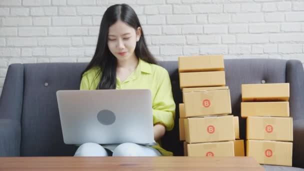 若いアジアの女性は オンラインショッピング ビジネス中小企業やスタートアップ 起業家や売り手とサービスの配信製品を購入する顧客の注文をチェックソファの上にラップトップコンピュータを使用して商人です — ストック動画