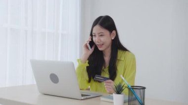 Genç Asyalı iş kadını akıllı telefondan bahsediyor ve evde dizüstü bilgisayarla online alışveriş yaparken kredi kartı tutuyor. Ödeme için banka kartı tutan kadın, iletişim konsepti..