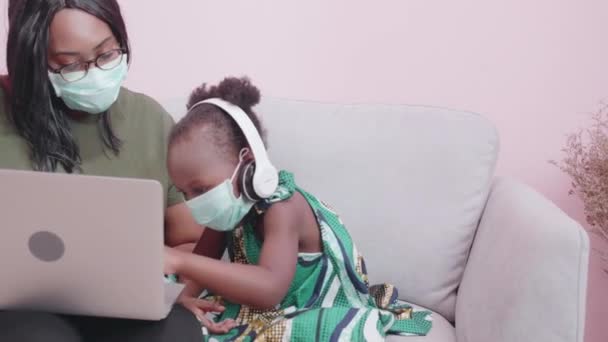 家で一緒に勉強し学ぶためにラップトップコンピュータを使用して顔のマスクを身に着けている若いアフリカの母親と娘 距離とEラーニングのためのヘッドフォンを身に着けている小さな子供 教育の概念 — ストック動画