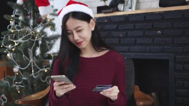 Genç Asyalı kadın Noel 'de kredi kartıyla online alışveriş yapıyor. Telefonla kadın satın alıyor ve ödemesini akıllı telefondan yapıyor. Noel' de xmas, mutlu yıllar, internetten alışveriş yapan müşteriler..