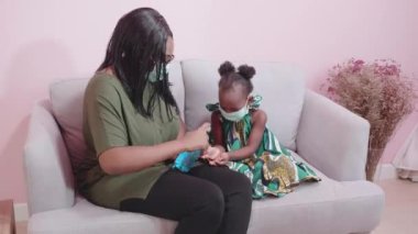 Afrikalı anne, yüz maskesi takan ve grip virüsünü korumak için dezenfektan kullanan ya da evdeki oturma odasında oturan anne ve çocuk, tıbbi maske takıyor ve el temizliği yapıyor..