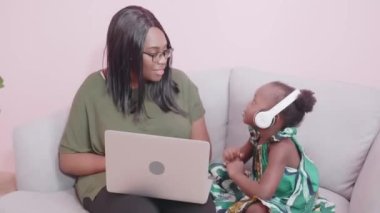 Genç Afrikalı anne ve kız müzik dinliyor ve evde rahatlamak için neşeli ve eğlenceli bir şekilde şarkı söylüyor, anne ve küçük çocuk dizüstü bilgisayar, aile ve eğitim konseptiyle e-öğrenim öğreniyor.