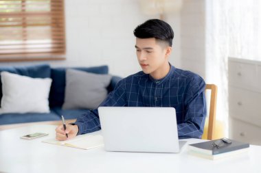 Genç Asyalı iş adamı evdeki dizüstü bilgisayarla çalışma ve kullanma planlarını deftere yazıyor. Finans, erkek çalışma ve öğrenme, iş ve iletişim konsepti hakkında notlar yazıyor..
