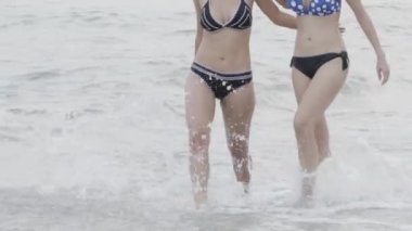 Güzel iki Asyalı kadın bikinili sahilde yürüyor ve su sıçratıyor, yaz tatilinde seksi bir kız tatil için seyahat ediyor, kadınlar tropikal ve yavaş çekimde denizde mayo giyiyor..