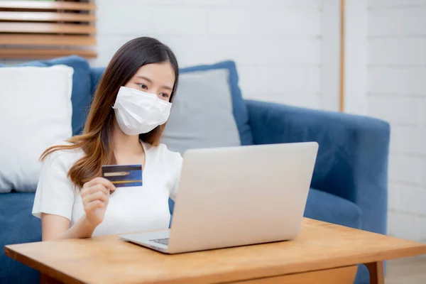 Young asian woman in face mask holding credit card shopping online with laptop computer buy and payment, girl using debit card purchase or transaction of finance, lifestyle and new normal concept.