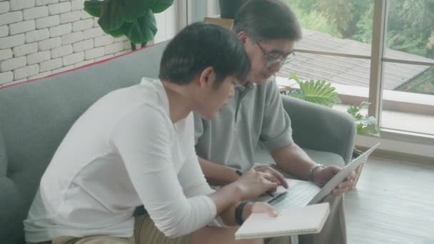 儿子坐在沙发上看笔记本电脑和写核对表的家庭在笔记本上与父亲 老人和年轻人在网上购物的财务费用 商业概念 — 图库视频影像