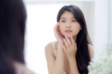 Genç ve güzel Asyalı bir kadın gülümsüyor cilt bakımı ve kozmetik ile cilt bakımını kontrol ederken aynaya bakıyor. Güzellik kızı, krem ve losyonla mutlu bir yüz ifadesi sergiliyor..