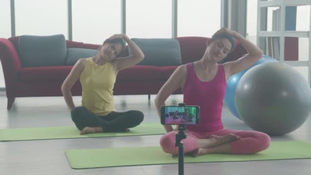 2人の若いアジアの女性がヨガで首を伸ばし 自宅でマットでスマートフォンでストリーミング ジムクラブ 健康とリラクゼーションのためのスポーツでアクティブな運動とオフィス症候群 — ストック動画