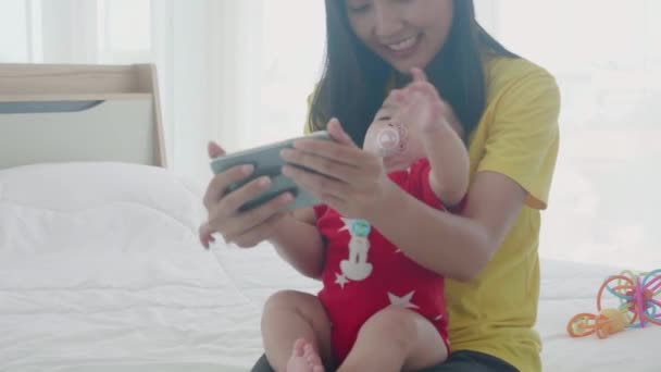 若いアジアの母親と娘のビデオ通話は インターネット上で教育や通信のためにスマートフォンを使用して 幸せな母親と赤ちゃんの女の子で一緒にベッドで家族と電話で — ストック動画