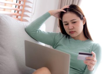 Kredi kartı ya da kredi kartı tutan genç Asyalı bir kadın, evdeki oturma odasındaki kanepede oturan kredi ve harcama stresiyle, bütçe ve finans konusunda endişeli ve hayal kırıklığına uğramış bir kadın, iş konsepti.