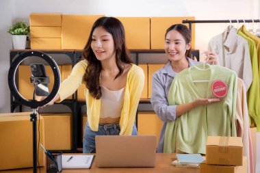 İki genç Asyalı kadın online kıyafet mağazasında canlı yayında canlı yayında sosyal güvenlik telefonuyla kıyafetlerini gösterirken, girişimci online kıyafet satışı ve sunumu için online satıcı,.
