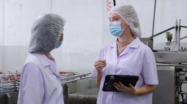Gıda işleme fabrikasında kalite kontrolü yapan müfettiş ekibi, üniformalı ve maskeli iki işçi kadın kalite ve tedarik zincirini incelemek için hijyen ve üretimi izlemek için tablet kullanıyor..
