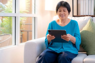 Kıdemli Asyalı kadın evdeki oturma odasında tablet okurken, internet bağlantısı ve dijital yaşam tarzı, sosyal medya için tablet izlerken, uzaktan öğrenirken, sohbet robotu veya yapay zeka kullanırken, kanepede dinleniyor..