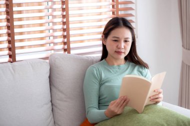 Genç Asyalı bir kadın, yüz ifadesiyle kitap okuyor. Oturma odasındaki koltukta oturuyor, kadın rahatlıyor ve kanepeye oturuyor. Edebiyat, etkinlik ve hobi, yaşam tarzı ve rahatlama okuyor..