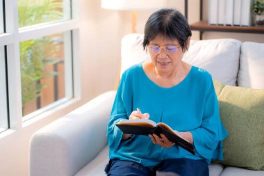 Asyalı son sınıf öğrencisi kadın kitap yazıyor. Oturma odasında dinleniyor. Yaşlı kadın, hafta sonu rahat, hobi ve eğlence dolu günlükler yazıyor..