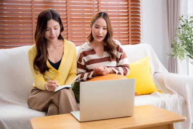 İki Asyalı kadın dizüstü bilgisayar görüntülerini kullanarak kanepede oturuyorlar. Ev ödevlerini birlikte yapıyorlar. Ev işleri için takım çalışması, ev idaresi, proje üzerinde işbirliği, serbest planlama..