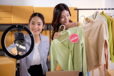 İki genç Asyalı kadın online kıyafet mağazasında canlı yayında canlı yayında sosyal güvenlik telefonuyla kıyafetlerini gösterirken, girişimci online kıyafet satışı ve sunumu için online satıcı,.