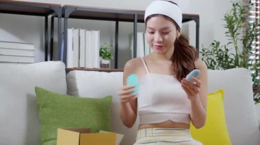 Kanepede oturan genç Asyalı kadın kozmetik kremi ya da losyon ile evdeki oturma odasında alışveriş, cilt bakımı, cilt bakımı, güzellik ve cilt sağlığı için cilt bakımı ve makyaj ürünü.