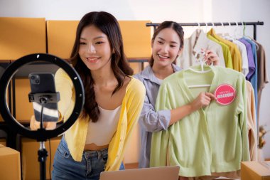 İki genç Asyalı kadın online kıyafet mağazasında canlı yayında canlı yayında sosyal güvenlik telefonuyla kıyafetlerini gösterirken, girişimci online kıyafet satışı ve sunumu için online satıcı,.