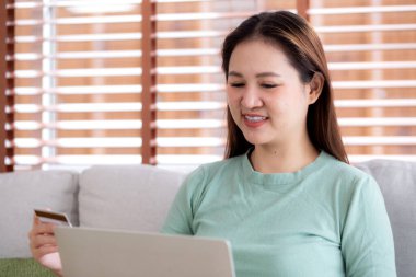 Kanepede oturan Asyalı genç bir kadın internet üzerinden dizüstü bilgisayar alışverişi yapıyor. İnternete kredi kartı alışverişi yapıyor. Kanepe, satın alma ve ödeme, iş ve yaşam tarzları ile kadın ödemesi yapıyor..