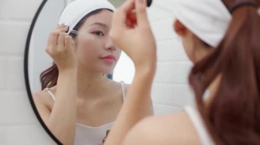 Güzel Asyalı genç bir kadın, yüz aynasına gençleştirme serumu uyguluyor. Güzellik kadını cilt, cilt bakımı, cilt bakımı, cilt bakımı, makyaj, kozmetik ve makyaj için kolajen uyguluyor..