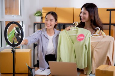İki genç Asyalı kadın online kıyafet mağazasında canlı yayında canlı yayında sosyal güvenlik telefonuyla kıyafetlerini gösterirken, girişimci online kıyafet satışı ve sunumu için online satıcı,.