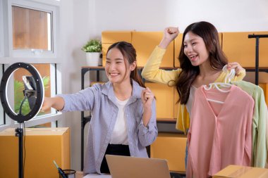 İki genç Asyalı kadın online kıyafet mağazasında canlı yayında canlı yayında sosyal güvenlik telefonuyla kıyafetlerini gösterirken, girişimci online kıyafet satışı ve sunumu için online satıcı,.