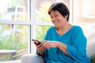 Üst düzey Asyalı kadın akıllı telefonuyla evdeki koltukta otururken gülümsüyor. Dijital yaşam tarzı, sosyal medyayla akıllı telefon kullanan yaşlı Asyalı. Sohbet robotu ya da yapay zeka..