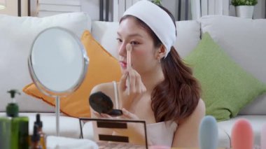 Güzel, mutlu, Asyalı bir kadın oturma odasında aynaya bakarak makyaj yapıyor. Güzel bir kadın makyaj yapıyor. Cazibe ve zarafet için yüz makyajı yapıyor..