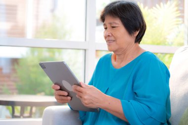 Kıdemli Asyalı kadın evdeki oturma odasında tablet okurken, internet bağlantısı ve dijital yaşam tarzı, sosyal medya için tablet izlerken, uzaktan öğrenirken, sohbet robotu veya yapay zeka kullanırken, kanepede dinleniyor..