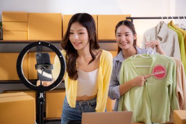 İki genç Asyalı kadın online kıyafet mağazasında canlı yayında canlı yayında sosyal güvenlik telefonuyla kıyafetlerini gösterirken, girişimci online kıyafet satışı ve sunumu için online satıcı,.