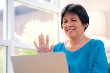 Üst düzey Asyalı kadın dizüstü bilgisayarında el sallıyor oturma odasındaki koltukta oturuyor, yaşlılara ait video kayıtları internette, kanepeye, emekliliğe ve teknolojiye uzaktan bakıyor..