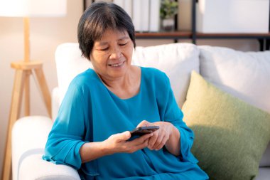 Üst düzey Asyalı kadın akıllı telefonuyla evdeki koltukta otururken gülümsüyor. Dijital yaşam tarzı, sosyal medyayla akıllı telefon kullanan yaşlı Asyalı. Sohbet robotu ya da yapay zeka..