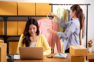 İki genç Asyalı iş kadını dizüstü bilgisayar, sevkiyat ve kargo için müşteri kıyafetlerini kontrol ediyor, çevrimiçi satıcı ve mağaza ile perakende satış, başlangıç veya küçük işletme, nakliye ve envanter.