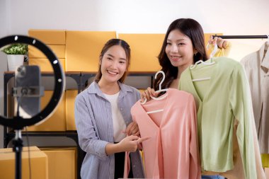 İki genç Asyalı kadın online kıyafet mağazasında canlı yayında canlı yayında sosyal güvenlik telefonuyla kıyafetlerini gösterirken, girişimci online kıyafet satışı ve sunumu için online satıcı,.