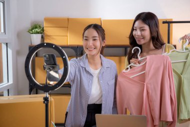 İki genç Asyalı kadın online kıyafet mağazasında canlı yayında canlı yayında sosyal güvenlik telefonuyla kıyafetlerini gösterirken, girişimci online kıyafet satışı ve sunumu için online satıcı,.