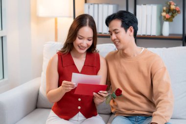 Sevgililer Günü 'nü romantik bir sürprizle birlikte koltukta oturan mutlu Asyalı çift evde gül ve kart okurken, aşkı hissederken, 14 Şubat ve yıldönümlerinde romantik.