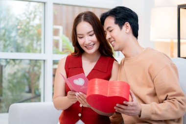 Asyalı genç adam ve kız arkadaş Sevgililer Günü 'nü kutluyorlar. Kartpostal ve hediye kutlamasıyla neşeli bir çift evde kanepede oturup tatil kutluyor ve aşkı hissediyorlar..