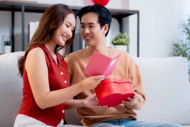 Asyalı genç adam ve kız arkadaş Sevgililer Günü 'nü kutluyorlar. Kartpostal ve hediye kutlamasıyla neşeli bir çift evde kanepede oturup tatil kutluyor ve aşkı hissediyorlar..