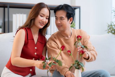 Mutlu Asyalı bir çift vazoda birlikte güller düzenliyor, evde romantik bir sevgililer günü aktivitesi, genç bir adam ve kadın kanepede oturuyor birbirlerine bağlanıyorlar ve aşk, romantik tatil ve ilişki.
