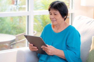 Kıdemli Asyalı kadın evdeki oturma odasında tablet okurken, internet bağlantısı ve dijital yaşam tarzı, sosyal medya için tablet izlerken, uzaktan öğrenirken, sohbet robotu veya yapay zeka kullanırken, kanepede dinleniyor..