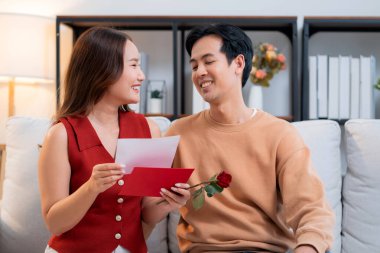 Sevgililer Günü 'nü romantik bir sürprizle birlikte koltukta oturan mutlu Asyalı çift evde gül ve kart okurken, aşkı hissederken, 14 Şubat ve yıldönümlerinde romantik.