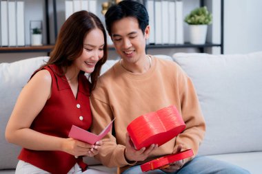 Asyalı genç adam ve kız arkadaş Sevgililer Günü 'nü kutluyorlar. Kartpostal ve hediye kutlamasıyla neşeli bir çift evde kanepede oturup tatil kutluyor ve aşkı hissediyorlar..