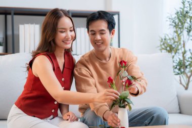 Mutlu Asyalı bir çift vazoda birlikte güller düzenliyor, evde romantik bir sevgililer günü aktivitesi, genç bir adam ve kadın kanepede oturuyor birbirlerine bağlanıyorlar ve aşk, romantik tatil ve ilişki.
